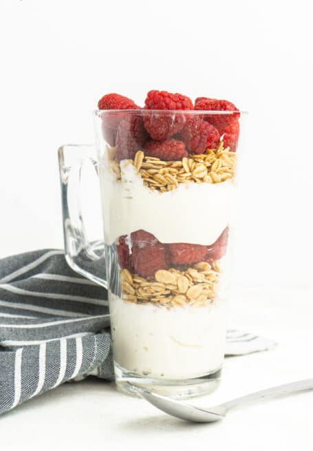 A tall glass mug layered with Greek yogurt, granola and fresh raspberries, with a spoon beside it.