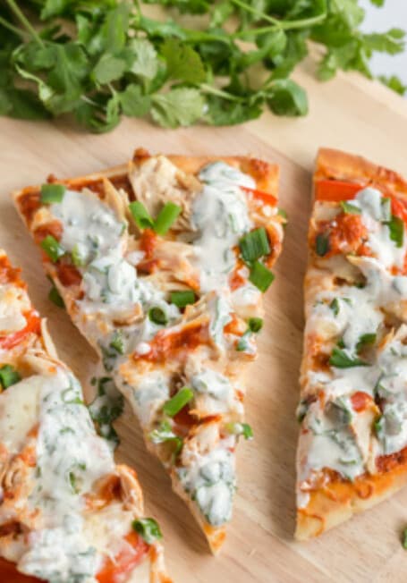 Slices of butter chicken flatbread pizza topped with shredded chicken, cheese, green onion and a creamy cilantro drizzle, with fresh cilantro behind.