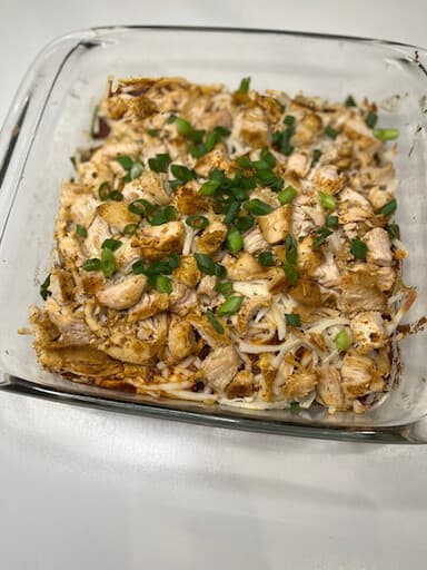 A glass baking dish of chicken enchilada lasagna topped with browned chicken, melted cheese and sliced green onions.