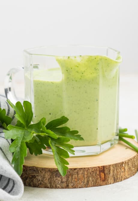 A glass jug of creamy green dressing on a wooden slice with fresh parsley and chives.