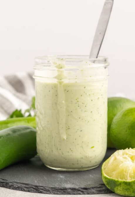 A jar of creamy jalapeno lime dressing with a spoon, beside fresh jalapenos and limes.