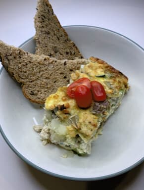 A slice of crustless quiche topped with cherry tomatoes in a shallow bowl, with two slices of seeded bread.