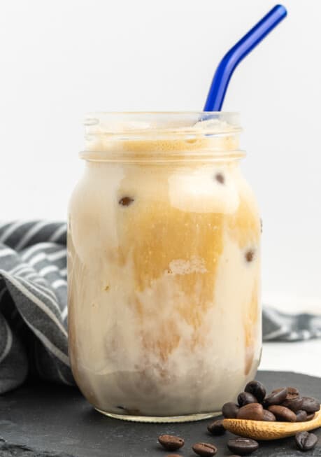 A glass mason jar of iced protein coffee with a blue straw, with coffee beans on a wooden spoon beside it.