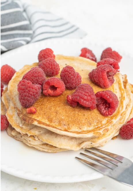 A stack of protein pancakes topped with fresh raspberries on a white plate.