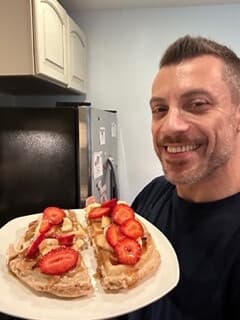 A smiling man holding a plate of protein waffles topped with peanut butter and sliced strawberries.