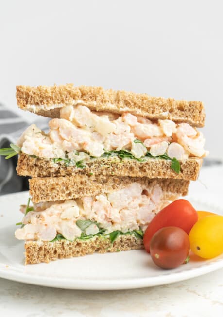 A stacked shrimp salad sandwich on whole grain bread with greens, beside cherry tomatoes on a white plate.