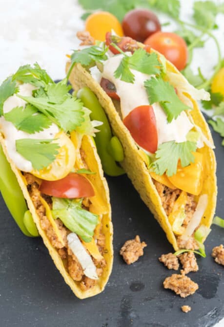Two crispy hard-shell turkey tacos topped with lettuce, tomato, cilantro and creamy sauce on a dark slate board.