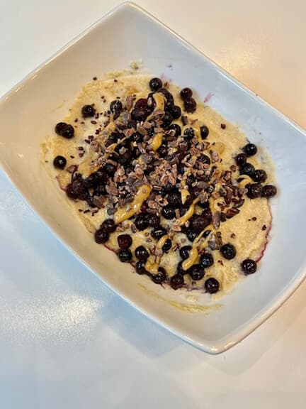 A white bowl of cream of rice topped with blueberries, nut butter drizzle and cacao nibs.